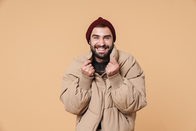 Портрет кавказского молодого человека в зимней куртке и шляпе, улыбаясь изолирован на бежевом