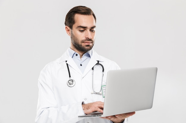 Portrait of caucasian young medical doctor with stethoscope working in clinic and holding laptop isolated over white wall