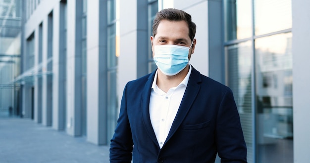 Photo portrait of caucasian young stylish businessman in medical mask looking at camera, smiling and standing at urban street. male outdoor in city at business center during coronavirus pandemic.