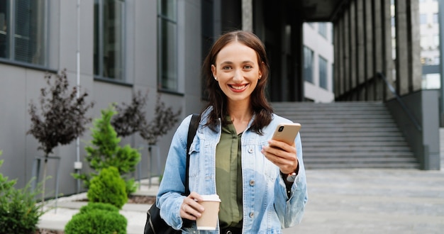 朝にスマートフォンとホットドリンクを保持し、カメラに笑顔の白人の若いスタイリッシュな女性の肖像画。コーヒーを飲む携帯電話を持つ美しい幸せな女子学生。屋外。