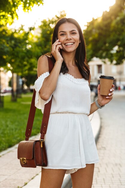 紙コップを持って屋外を歩きながらスマートフォンで話している夏の服を着た白人の若い女性の肖像画