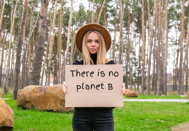 Photo portrait of caucasian young woman in hat holding cardboard with text outdoors nature background