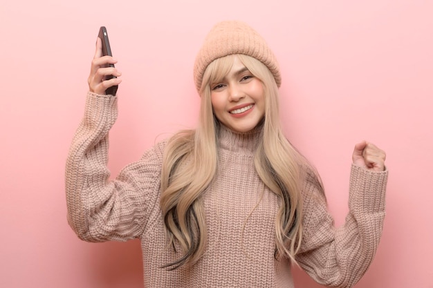 Portrait of Caucasian young woman wearing sweater holding smart phone over pink background