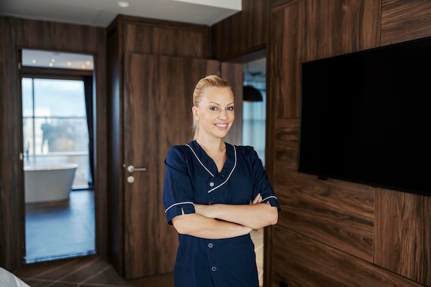 Portrait of a chambermaid standing in a hotel room with arms crossed and smiling at the camera