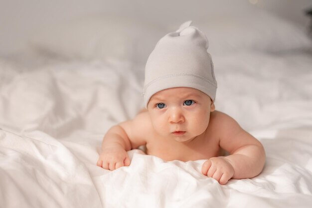 Portrait of a charming baby with blue eyes in a white cap lying on his tummy on white bed linen