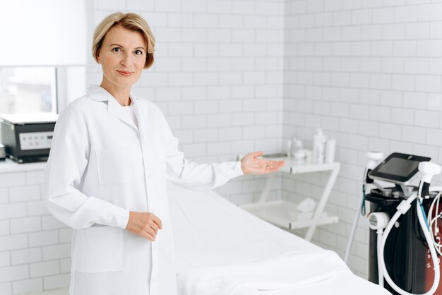 Portrait of charming beautician in white lab coat looking at camera and smiling while standing near daybed. Stock photo