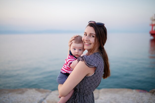 Portrait of Charming brunette holding her adorable 6 months old son while standing on the beach at sunset.