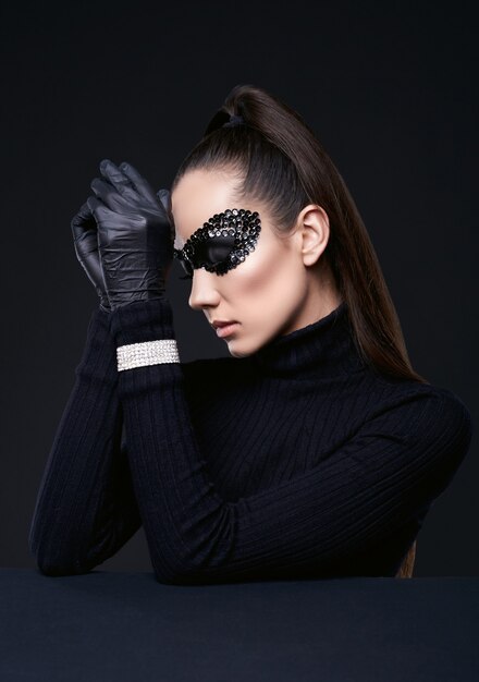 Portrait of charming elegant brunette woman in turtleneck sweater and sequins mask posing on black in studio