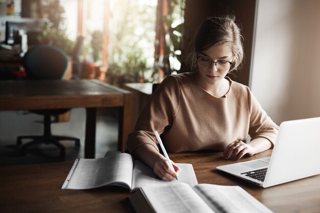 Portrait of charming focused caucasian female student in glasses, writing with pen in notebook, working with laptop, gathering information from internet.