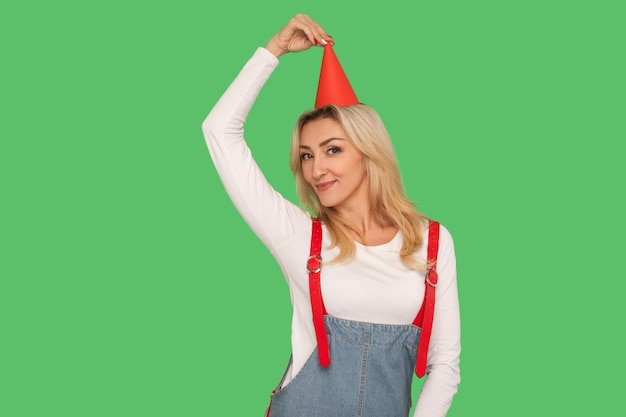 Portrait of charming happy adult woman in stylish overalls touching party cone on her head and looking at camera with smile celebrating birthday indoor studio shot isolated on green background