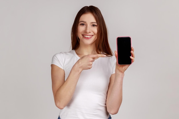 Portrait of charming happy woman pointing at cellphone advertising gadget and online application good cellular tariffs plan wearing white Tshirt Indoor studio shot isolated on gray background