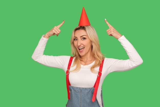 Portrait of charming joyful adult woman in stylish overalls pointing at party cone on her head and looking at camera with toothy smile celebrating birthday studio shot isolated on green background