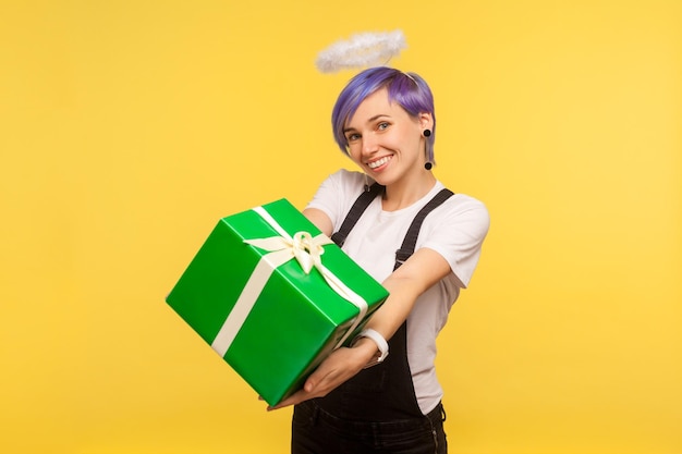 Portrait of charming joyful hipster girl with angelic halo over violet short hair giving wrapped present box to camera congratulating and sharing gift isolated on yellow background studio shot