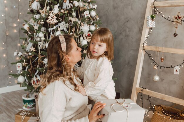 크리스마스 트리 근처에 앉아 사랑하는 어머니로부터 선물을 받는 매력적인 어린 소녀의 초상화. 아름 다운 여자는 그녀의 아이에 게 새 해 선물을 만든다.