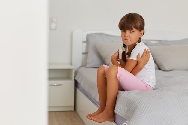 Portrait of charming offended sad female kid wearing white t shirt and pink short, looking at camera, crying, feeling sorrow, posing on bed in light room alone.