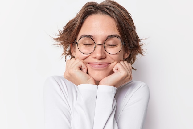Portrait of charming pleased brunette woman keeps hands under chin closes eyes has dreamy expression recalls something in mind wears rounded spectacles casual jumper isolated over white background