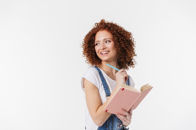 Portrait of charming redhead curly woman 20s wearing denim jumpsuit smiling and writing notes in her diary or day planner isolated over white wall