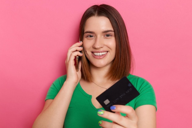 Ritratto di affascinante donna caucasica soddisfatta che indossa abiti in stile casual in piedi isolato su sfondo rosa in possesso di carta di credito parlando al cellulare guardando la fotocamera con un sorriso