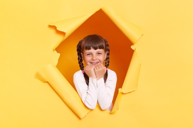 Portrait of charming satisfied little girl with braids wearing white turtleneck posing in torn hole of yellow paper wall keeping fists under chin looking at camera with happy expression