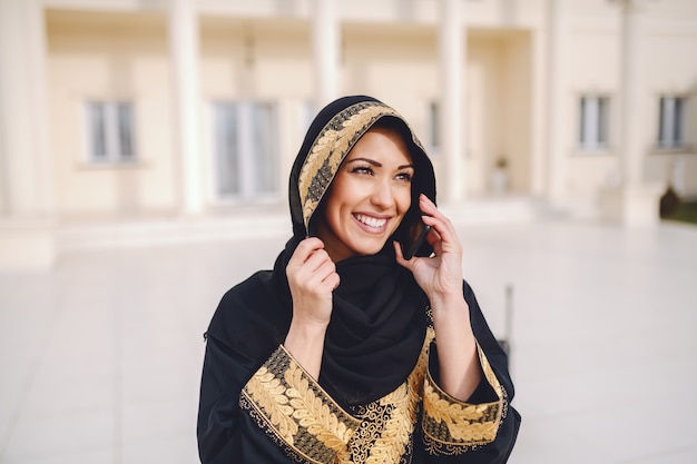 Портрет очаровательной усмехаясь мусульманской женщины одел в традиционной носке используя умный телефон пока стоящ снаружи.