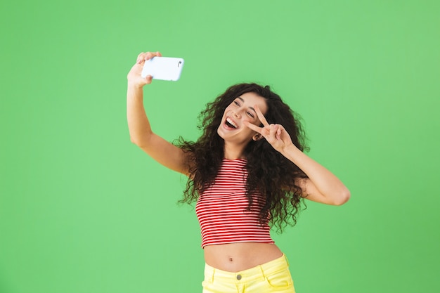 Portrait of charming woman 20s wearing summer clothes smiling and using cell phone for selfie isolated on green