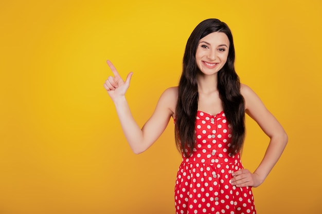 Portrait of charming woman finger indicate up empty space wear dotted dress on yellow background