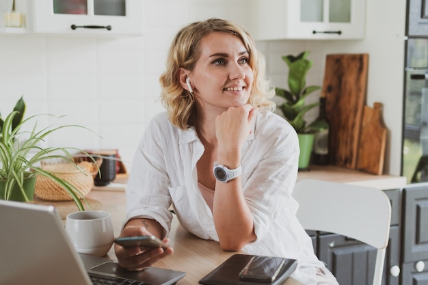 Portrait of charming young woman in headphones with laptop working at home in the kitchen