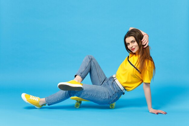 스튜디오의 파란색 벽 배경에 격리된 노란색 스케이트보드와 함께 카메라를 바라보고 앉아 있는 생생한 캐주얼 옷을 입은 매력적인 젊은 여성의 초상화. 사람들이 라이프 스타일 개념입니다. 복사 공간을 비웃습니다.