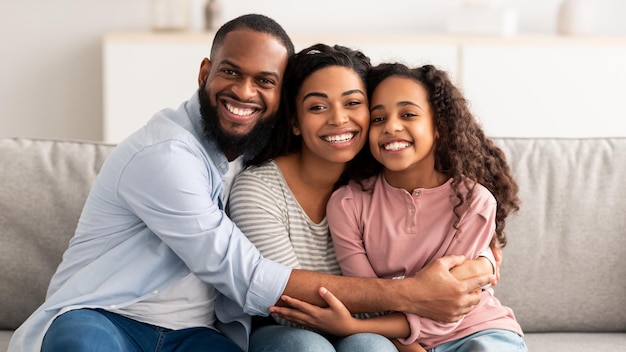 Portrait of cheerful African American family of three hugging sitting on the couch at home, posing for photo and looking at camera. Smiling young bearded man embracing his loving wife and daughter