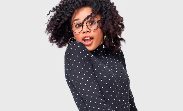 Portrait of cheerful African American woman smiling and spinning with her healthy hair feeling happy posing over white wall Beautiful Afro female blowing hair wearing transparent eyeglasses