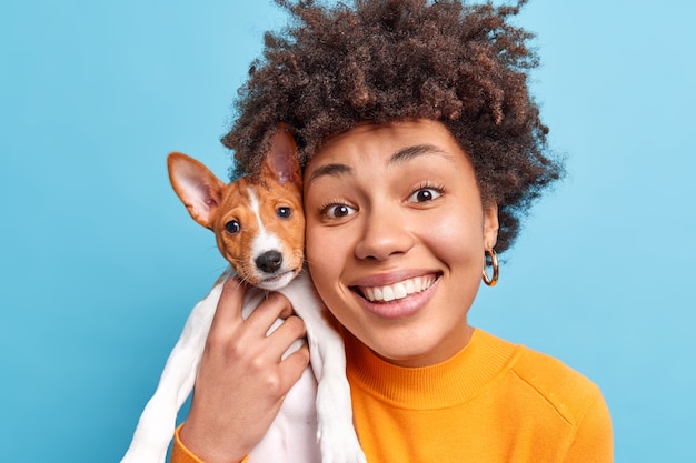 Portrait of cheerful Afro American woman dog owner keeps small pedigree puppy closely to face happy to get pet as present has new friends looks happily  isolated over blue wall