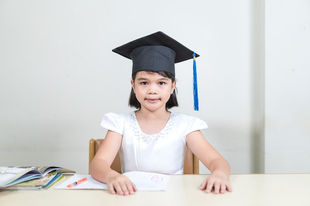 Portrait of cheerful asian little girl student wear a\
mortarboard or graduation hat hold pencil and write on notebook\
with white background with copy space. education graduation concept\
stock photo