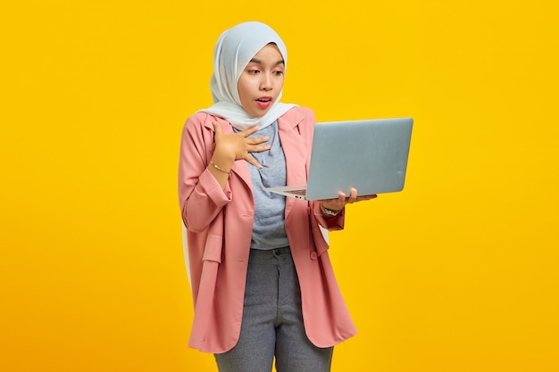 Portrait of a cheerful asian woman holding laptop and\
celebrating success isolated over yellow background