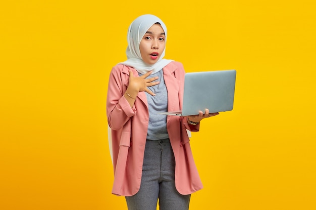 Portrait of a cheerful asian woman holding laptop and celebrating success isolated over yellow background