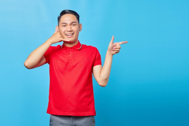 Portrait of cheerful Asian young man doing phone sign gesture and pointing finger at copy space isolated on blue background