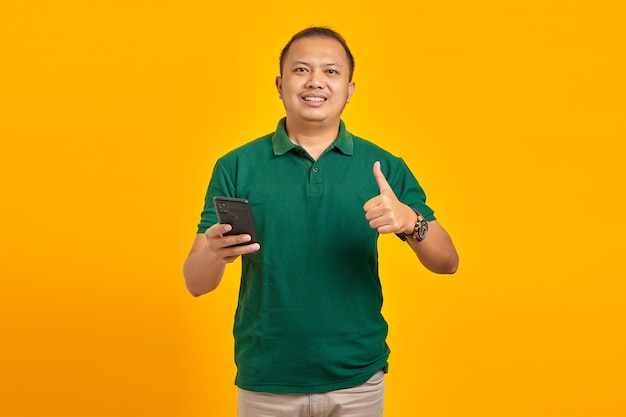 Portrait of cheerful Asian young man holding mobile phone and showing thumbs up sign