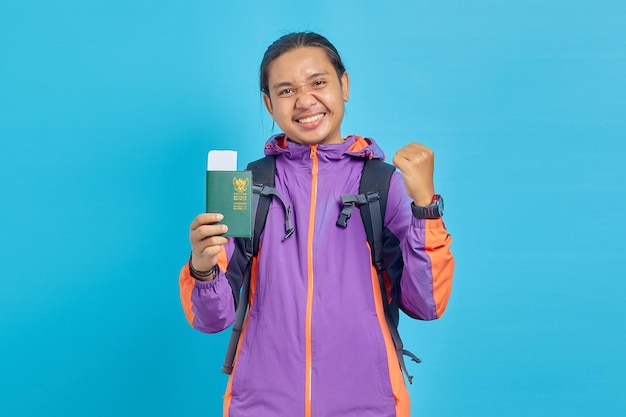 Portrait of cheerful asian young man showing parport book and making successful gesture isolated on blue background