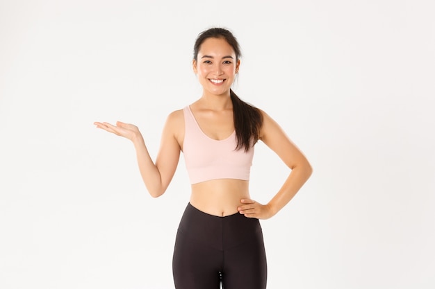   Portrait of cheerful attractive asian fitness coach, female athlete introduce or demonstrate product for workout, holding something on hand.