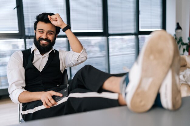 Portrait of cheerful bearded businessman sitting at office desk with feet up on table with laptop finished work job done completed all tasks