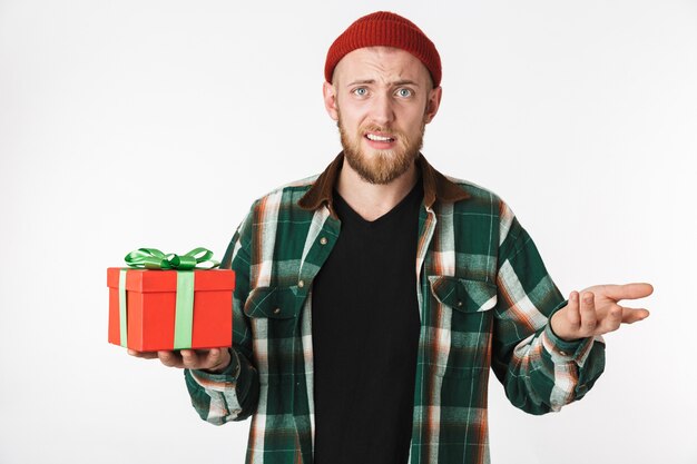 Portrait of cheerful bearded guy wearing hat and plaid shirt holding present box, while standing isolated over white background