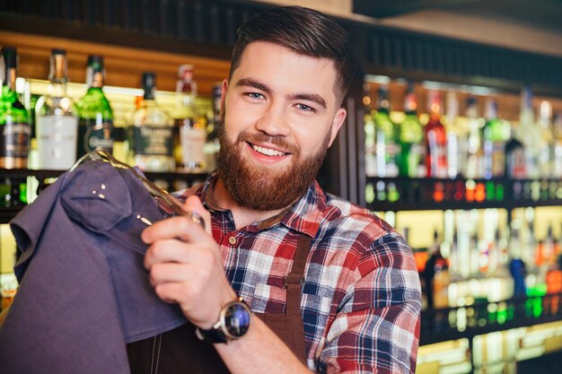 Portrait of cheerful bearded young barman wiping glasses in bar