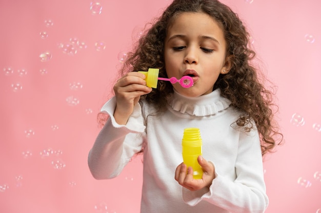 Портрет веселой красивой девочки, пускающей мыльные пузыри на розовом фоне с копировальным пространством Великолепный ребенок, играющий с мыльными пузырями Детский отдых и концепция дня рождения