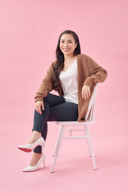 Portrait of a cheerful beautiful girl sitting on a chair and looking away isolated over pink background