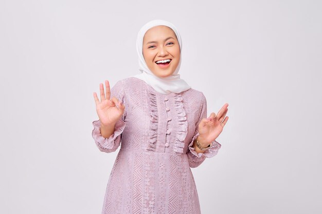 Портрет веселой красивой молодой азиатской мусульманки в хиджабе и фиолетовом платье, показывающей знак "хорошо" на белом фоне студии