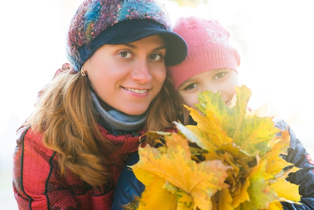 Portrait of a cheerful beautiful young mother with her pretty daughter holding autumn maple yellow leaves in her hands while walking in the park. Concept family ties and traditions