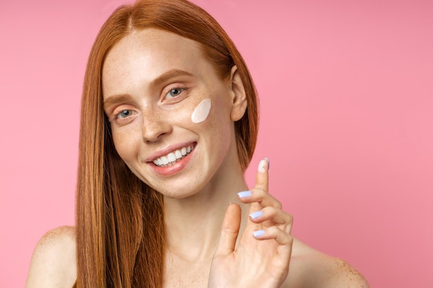 Foto il ritratto di una modella di bellezza allegra ha una pelle fresca e pura, applica una crema antirughe, sorride ampiamente con i denti bianchi, guardando con espressione felice su sfondo rosa. prodotto di bellezza, cura della pelle, benessere.