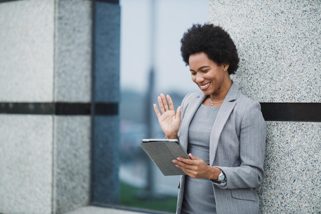 Портрет веселой темнокожей деловой женщины, делающей видеозвонок на цифровом планшете перед корпоративным зданием.