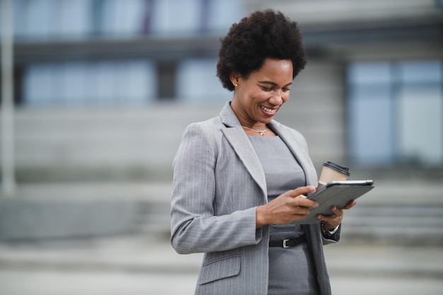 Portrait of a cheerful black business woman surfinh internet on a digital tablet while having a coffee break in front a corporate building.