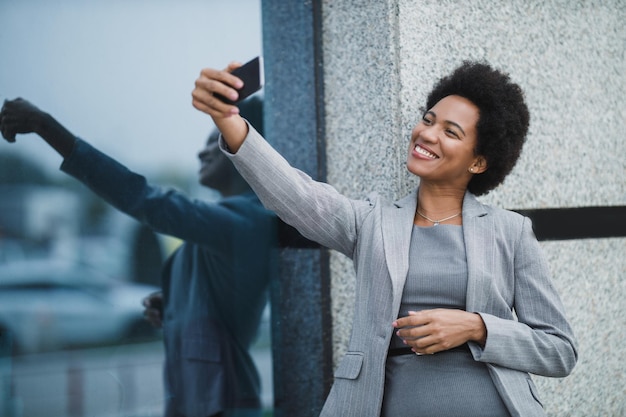 Ritratto di una donna d'affari nera allegra che scatta foto con il suo smartphone durante una breve pausa davanti a un edificio aziendale.