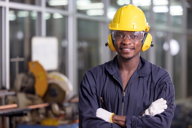 Portrait of cheerful black worker wearing protective headphones posing and enjoying work at background factory